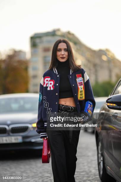 Mary Leest wears silver earrings, a black turtleneck / cropped top, a navy blue felt with embroidered red / yellow / blue print pattern teddy coat,...