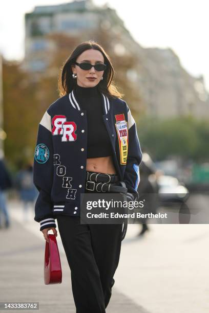 Mary Leest wears silver earrings, a black turtleneck / cropped top, a navy blue felt with embroidered red / yellow / blue print pattern teddy coat,...