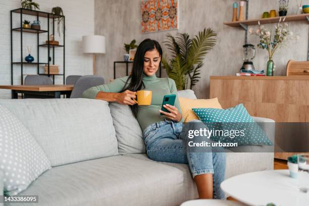 mujer joven tomando café y enviando mensajes de texto por teléfono en casa - chica-tomando-cafe fotografías e imágenes de stock