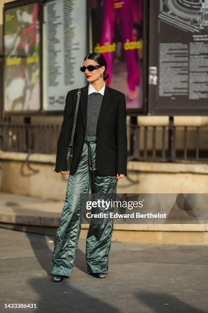 Zoia Mossour wears black sunglasses from Prada, silver earrings, a white shirt, a dark gray wool pullover, a black blazer jacket, a khaki grained...