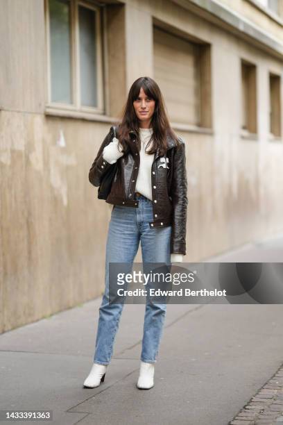 Astrid Boutrot wears a white latte wool pullover, a dark brown shiny varnished leather jacket from Courreges, blue faded denim large pants, white...