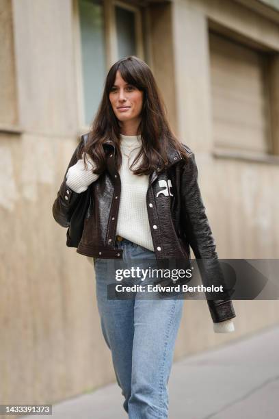 Astrid Boutrot wears a white latte wool pullover, a dark brown shiny varnished leather jacket from Courreges, blue faded denim large pants, a black...