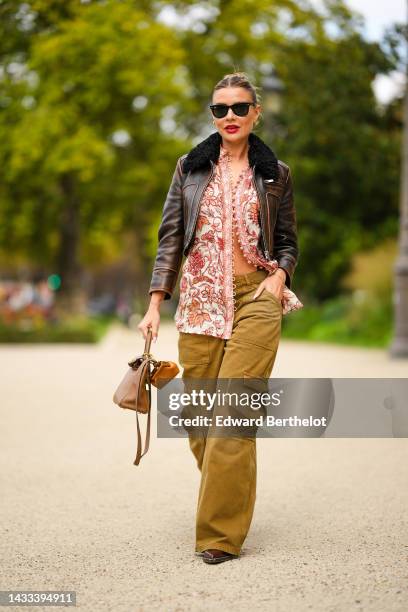 Veronica Ferraro wears black sunglasses from Ray Ban, gold earrings, a beige with red and orange flower print pattern buttoned blouse, a dark brown...