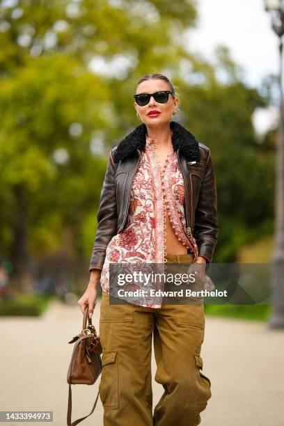 Veronica Ferraro wears black sunglasses from Ray Ban, gold earrings, a beige with red and orange flower print pattern buttoned blouse, a dark brown...