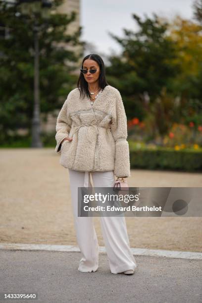 Sai De Silva wears black cat eyes sunglasses, gold earrings, a white pearls and gold large chain necklace, a white latte sheep oversized belted...