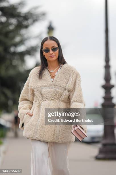 Sai De Silva wears black cat eyes sunglasses, gold earrings, a white pearls and gold large chain necklace, a white latte sheep oversized belted...