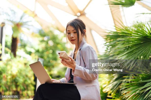 confident young asian businesswoman using smart phone while working on laptop in a modern greenhouse - young agronomist stock pictures, royalty-free photos & images