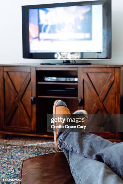 man relaxes in front of tv with feet on ottoman - pies cruzados en los tobillos fotografías e imágenes de stock