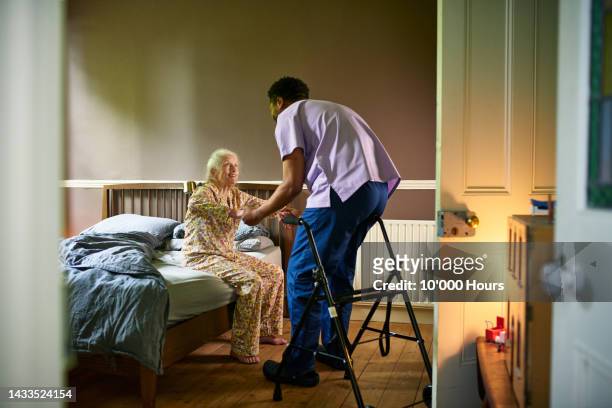 young male care worker helping woman off bed with walking frame nearby - residencia de ancianos fotografías e imágenes de stock