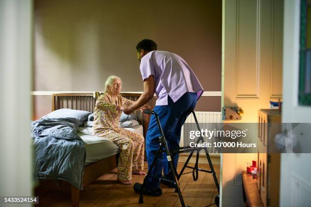 young male care worker helping woman off bed with walking frame nearby - seniorenhuis stockfoto's en -beelden