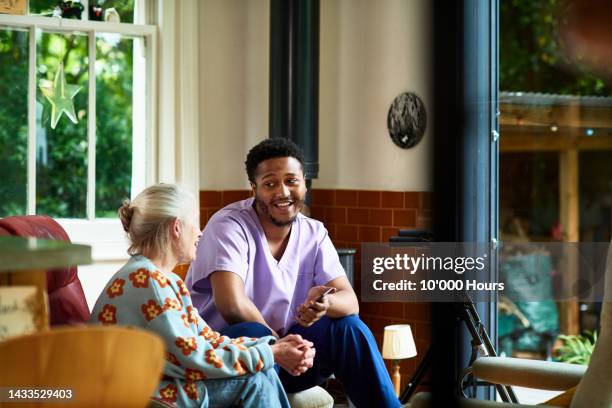 cheerful young black male care worker holding smartphone and chatting to senior woman - encouragement stock pictures, royalty-free photos & images
