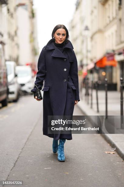 Mary Leest wears silver earrings, a black turtleneck wool pullover, a navy blue wool long oversized coat, a black shiny leather handbag, blue velvet...