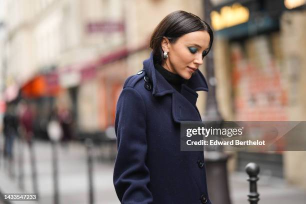 Mary Leest wears silver earrings, a black turtleneck wool pullover, a navy blue wool long oversized coat, a black shiny leather handbag, outside...