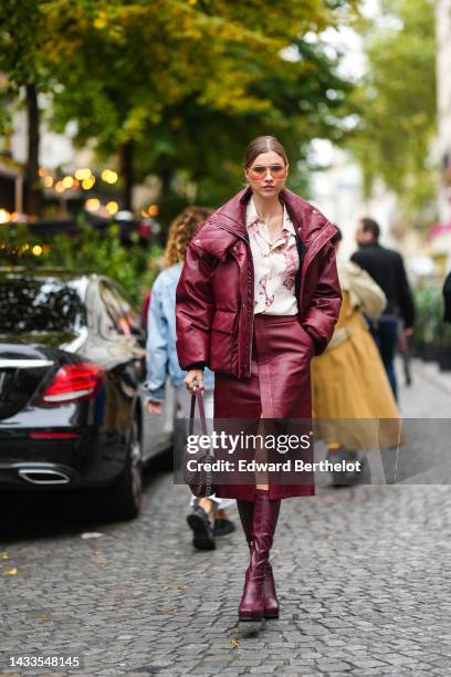 Zita D'Hauteville wears brown sunglasses, gold pendant earrings, a gold chain necklace, a burgundy shiny leather oversized puffer jacket, a white...