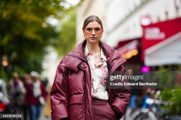 Zita D'Hauteville wears brown sunglasses, gold pendant earrings, a gold chain necklace, a burgundy shiny leather oversized puffer jacket, a white...