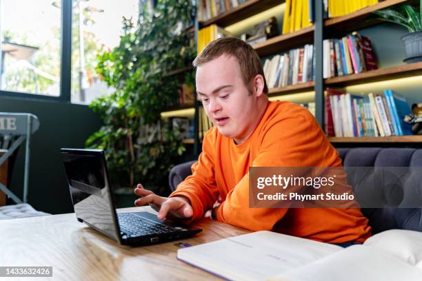 concentrated young man with down syndrome sitting and studying indoors at university, using laptop - disability computer stock pictures, royalty-free photos & images