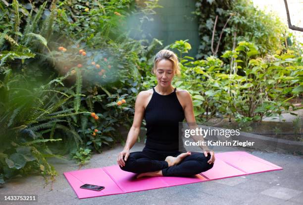 woman meditating in the garden, sitting cross-legged on exercise mat - yoga foto e immagini stock