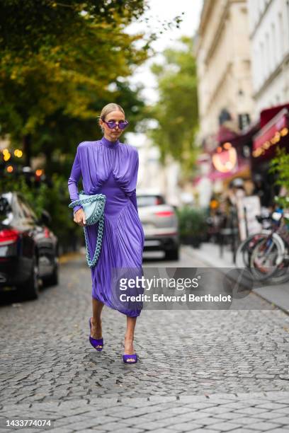 Leonie Hanne wears purple cat eyes sunglasses, silver earrings, a purple turtleneck / long sleeves / wrap pleated midi dress, a pale blue matte...