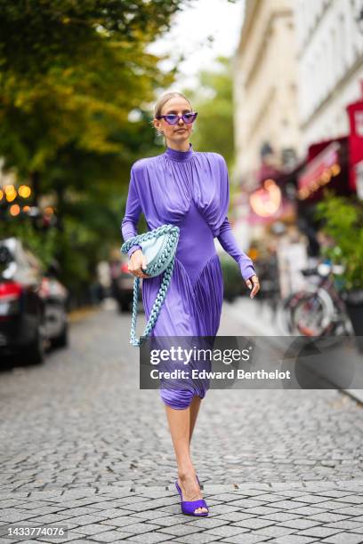 Leonie Hanne wears purple cat eyes sunglasses, silver earrings, a purple turtleneck / long sleeves / wrap pleated midi dress, a pale blue matte...