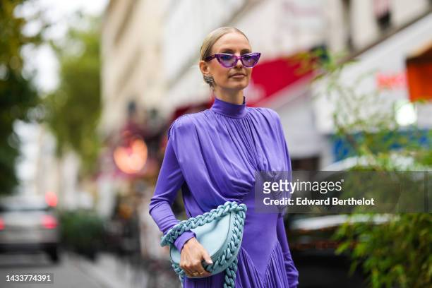 Leonie Hanne wears purple cat eyes sunglasses, silver earrings, a purple turtleneck / long sleeves / wrap pleated midi dress, a pale blue matte...