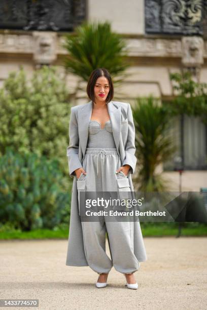 Tiffany Hsu wears a pale gray long wool coat, a pale gray heart-neck / shoulder-off body top, pale gray cargo sport pants, white shiny leather...
