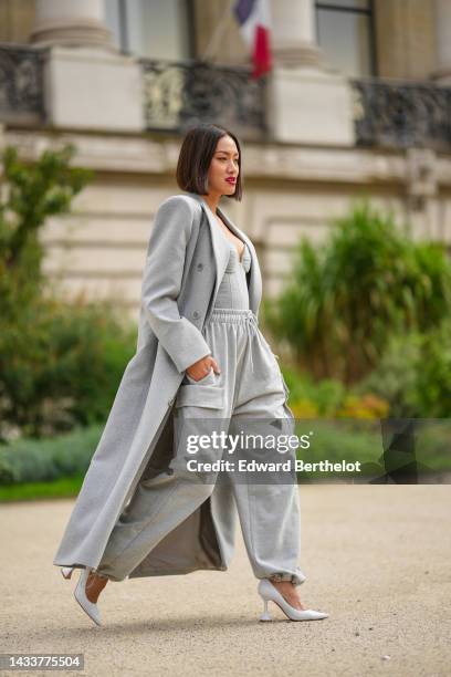 Tiffany Hsu wears a pale gray long wool coat, a pale gray heart-neck / shoulder-off body top, pale gray cargo sport pants, white shiny leather...