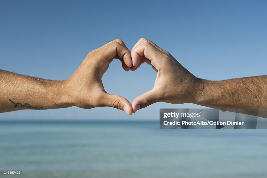 Two men's hands forming heart shape by ocean, cropped