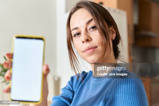 close-up of a young woman sitting holding smartphone showing your purchase online at home while looking at camera against blurred kitchen background - woman portrait kitchen laptop bildbanksfoton och bilder