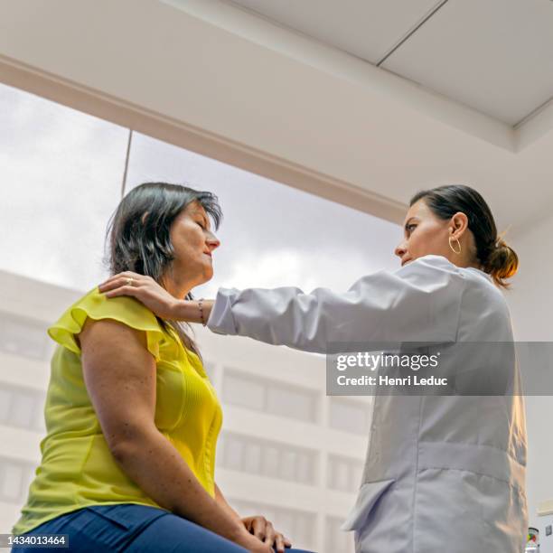 low-angle of female latin doctor holding hand on shoulder of sitting latin woman patient with close eyes - mature latin women fotografías e imágenes de stock