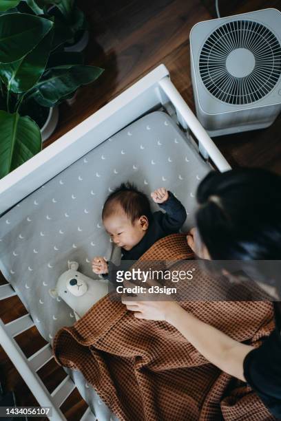 high angle shot of loving young asian mother taking care and tucking in her newborn baby daughter into crib in the nursery. love, care and tenderness. motherhood, parenthood concept - chambre nouveau né famille photos et images de collection
