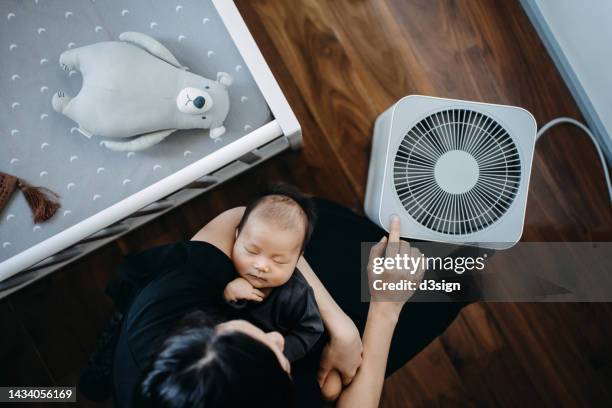 high angle shot of asian mother turning on home air purifier while carrying her sleeping newborn baby daughter in arms in the nursery. fresh air. cleaning and removing dust and bacteria. healthier life and living concept - air purifier fotografías e imágenes de stock