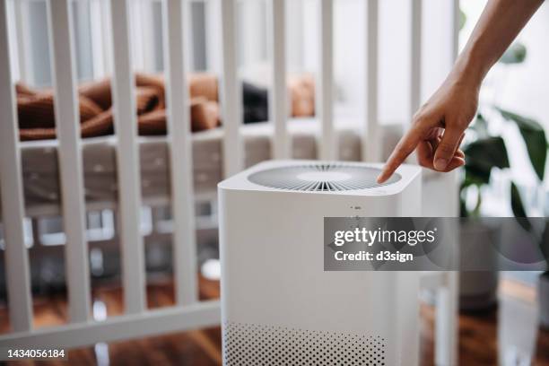 close up hand of a mother turning on home air purifier for her newborn baby who is sleeping in the crib in the nursery. fresh air. cleaning and removing dust and bacteria. healthier life and living concept - air purifier fotografías e imágenes de stock