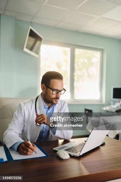male doctor taking notes while working on laptop in the office. - doctor using laptop stock pictures, royalty-free photos & images