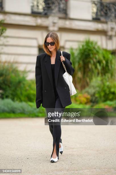Charlotte Groeneveld wears black sunglasses, a black shoulder-off / tank-top, black legging slit / split pants, a black oversized blazer jacket, a...