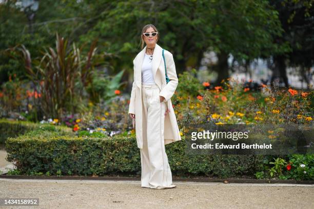 Darja Baranik wears a beige plastic cat eyes sunglasses, gold earrings, a black leather necklace with silver heart pendant, a white tank-top, a white...