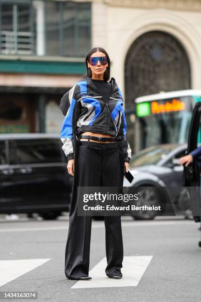 Julia Pelipas wears black large futurist sunglasses, a black cropped top, a black / pale gray / royal blue striped print pattern zipper jacket, a...