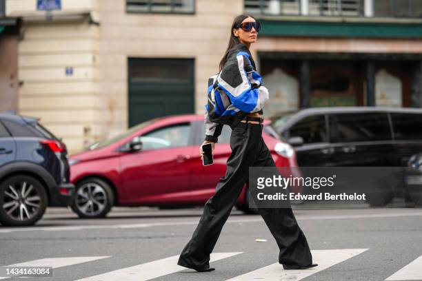 Julia Pelipas wears black large futurist sunglasses, a black cropped top, a black / pale gray / royal blue striped print pattern zipper jacket, a...
