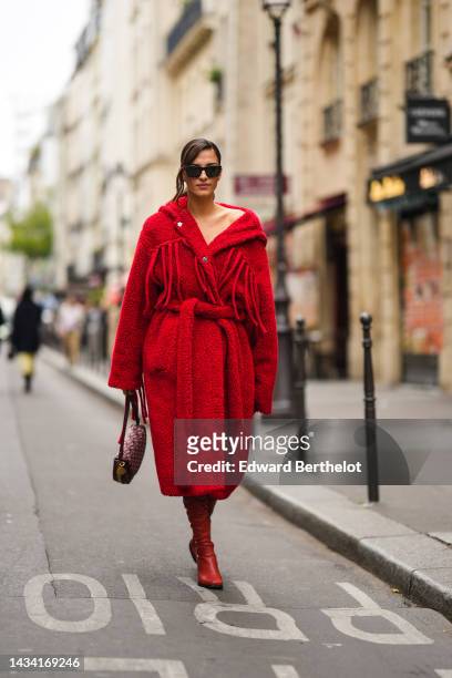 Gili Biegun wears black sunglasses, a red fluffy / sheep hoodie / oversized belted long coat, red shiny leather pointed / block heels knees boots /...