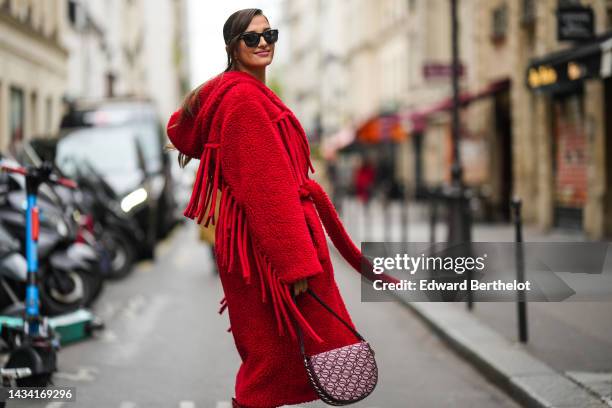 Gili Biegun wears black sunglasses, a red fluffy / sheep hoodie / oversized belted long coat, a pink and burgundy print pattern fabric handbag ,...