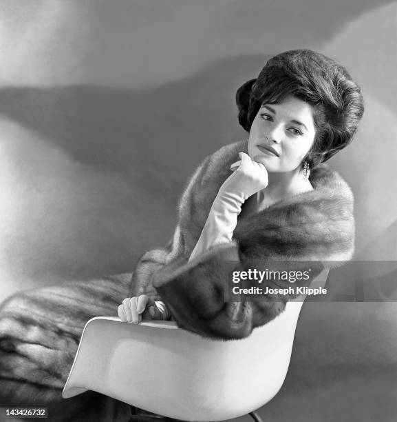 Woman models a mink coat as she sits in a molded plastic chair and rests her chin on her gloved hand, Washington DC, 1961.