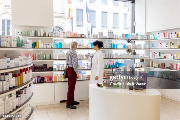 woman pharmacist assisting senior customer at pharmacy shop - farmacia fotografías e imágenes de stock