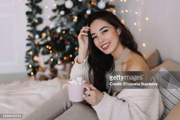 a latin american smiling woman in knitted pajamas holds a mug of hot coffee with marshmallows resting lying on the bed on christmas eve in a decorated room with a christmas tree at the indoor house - korean mexican woman stock pictures, royalty-free photos & images