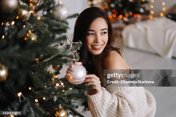 a beautiful multiracial asian woman in a knitted cardigan relaxes, smiles and laughs in a cozy room decorated with christmas decor and a christmas tree at home. cheerful girl of mixed race brunette having fun in the apartment on the new year's holiday - year long stock pictures, royalty-free photos & images