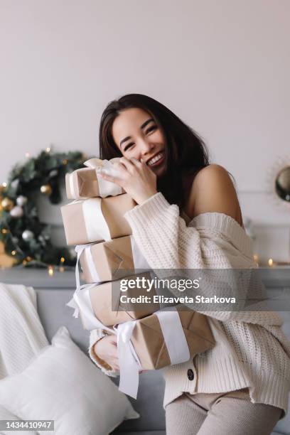 funny cheerful asian young woman in knitted home clothes holds a lot of christmas gifts and gift boxes preparing for the holiday in december in a decorated room at home - korean mexican woman stock pictures, royalty-free photos & images