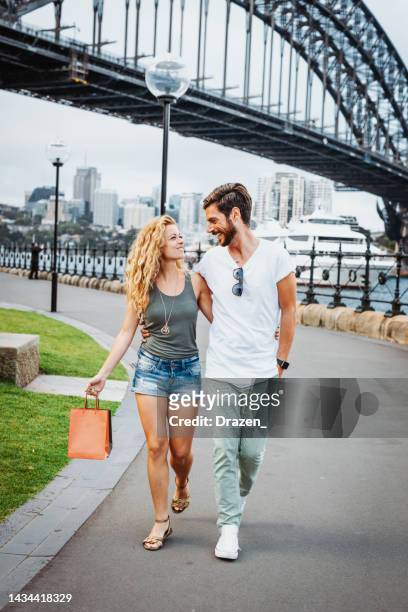 millennial couple walking with shopping bags in sydney, near sydney harbour bridge. christmas gifts for friends and family - sydney christmas lights 2017 stock pictures, royalty-free photos & images