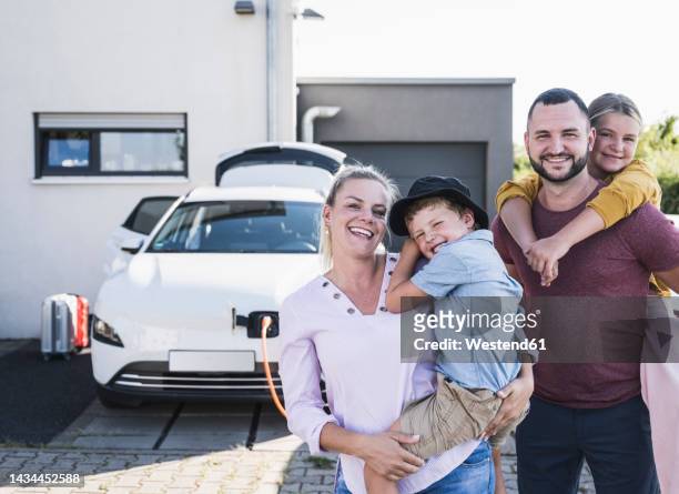 happy family standing in front of family home and charging electric car - familie unterwegs stock-fotos und bilder