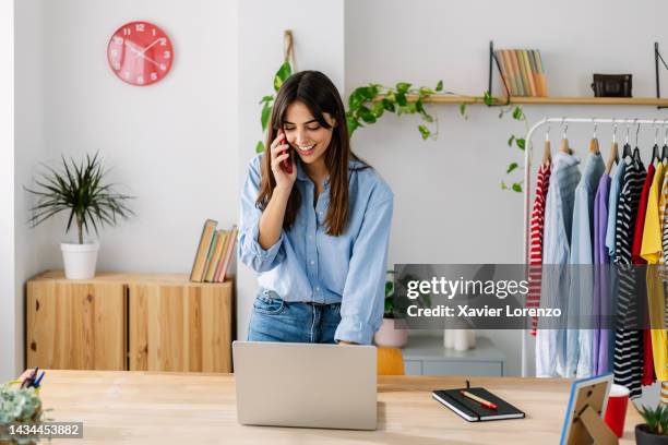 happy young adult small business female owner talking on mobile phone while working on laptop computer in clothing store - sarta foto e immagini stock