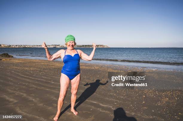 smiling elderly woman flexing muscles at beach - old woman in swimsuit stock-fotos und bilder