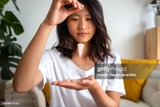 young asian woman using rose quartz pendulum at home. divination tool for spiritual practice. spirituality - fortune telling stock pictures, royalty-free photos & images