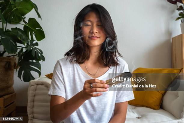 young asian woman holding a smoking palo santo smudging herself and the room. spirituality and meditation - fumigation photos et images de collection